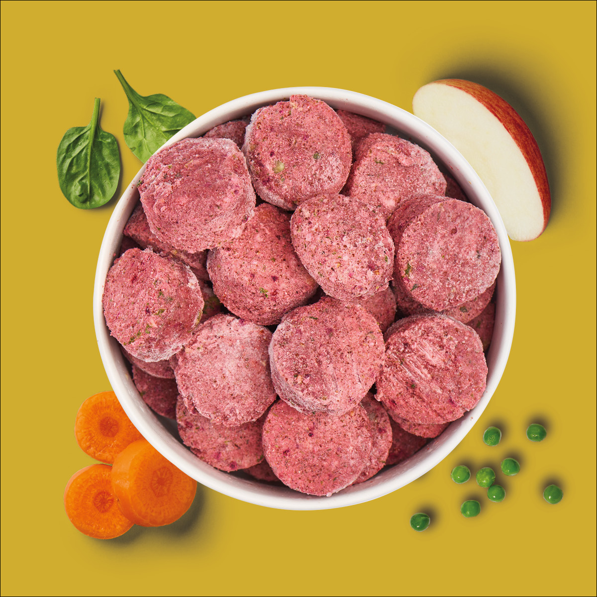 Nutriment BARF In One BUFFY Insect Weiße Schüssel mit gefrorenen roten Kroketten, umgeben von Spinat, Apfel, Karotte und Erbsen auf gelbem Hintergrund